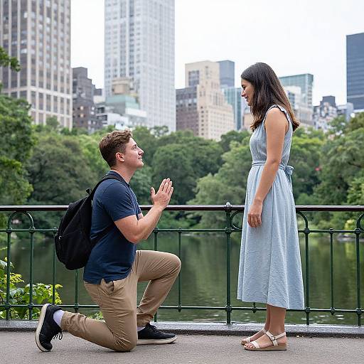 Photograph: Man kneeling, hands raised, proposing to woman in blue dress on city park overlook with skyscrapers and trees in background.