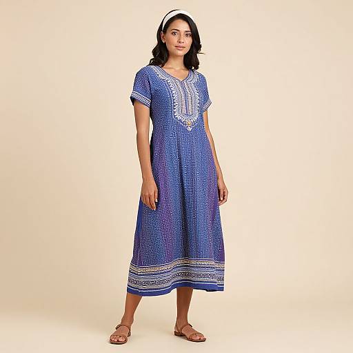 Photograph of a young woman with medium brown skin, wearing a blue, embroidered, ankle-length dress, white headband, and brown sandals, standing