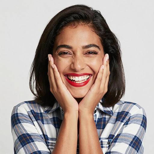 Photograph of a smiling young woman with dark hair, red lipstick, and white teeth, wearing a plaid shirt, hands on cheeks against a plain