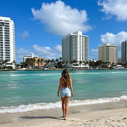 Photograph of a woman with long brown hair in a blue bikini top and white skirt, standing on a sandy beach, facing turquoise ocean with tall white