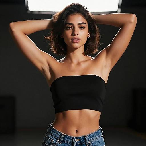 Photograph of a young woman with light brown skin and dark brown, shoulder-length hair, wearing a black strapless crop top and blue jeans, standing