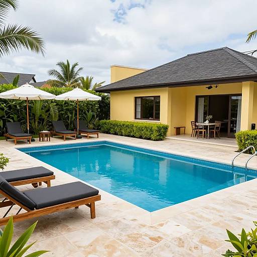 Photograph of a sunny backyard with a rectangular blue pool, yellow house, black-roofed patio, white umbrellas, black lounge chairs, and