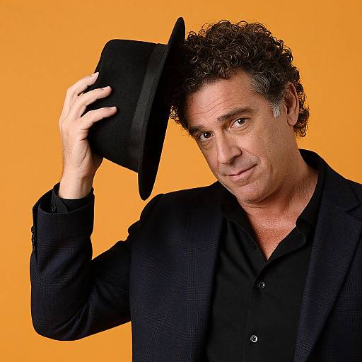 Photograph of a middle-aged man with curly brown hair, wearing a black hat, black button-up shirt, and black blazer, smiling against a