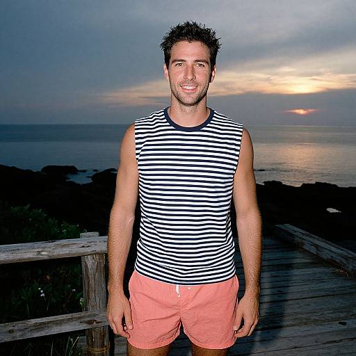 Photograph of a smiling man with short dark hair, wearing a black-and-white striped tank top and coral shorts, standing on a wooden boardwalk at