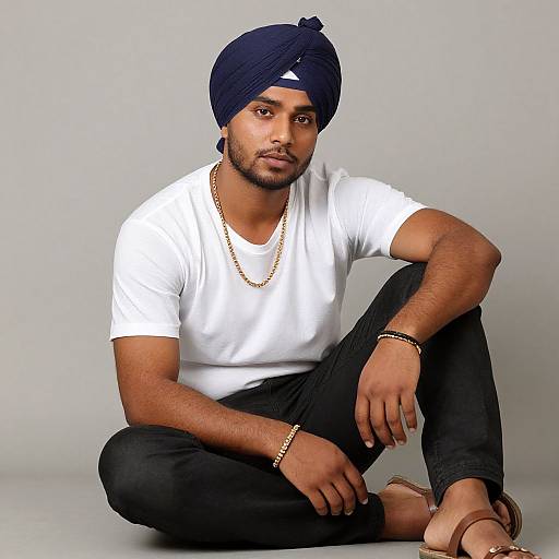 Photograph of a South Asian man with dark skin, black turban, white t-shirt, black pants, gold chain, bracelet, sitting cross-legged