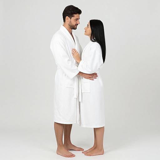 Photograph of a couple in white bathrobes standing barefoot, facing each other closely, with the man holding the woman's waist and her hands