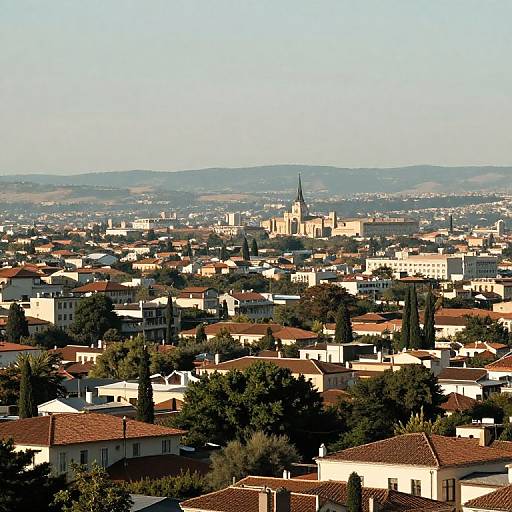 Photograph of a sunlit Mediterranean town with red-tiled roofs, white buildings, a prominent church with a tall spire, and lush green trees