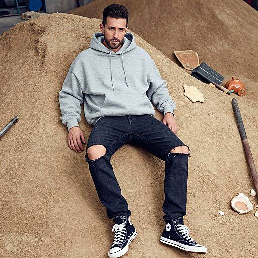 Photograph of a bearded man with short dark hair, wearing a gray hoodie, black ripped jeans, and black sneakers, sitting on a sand pile