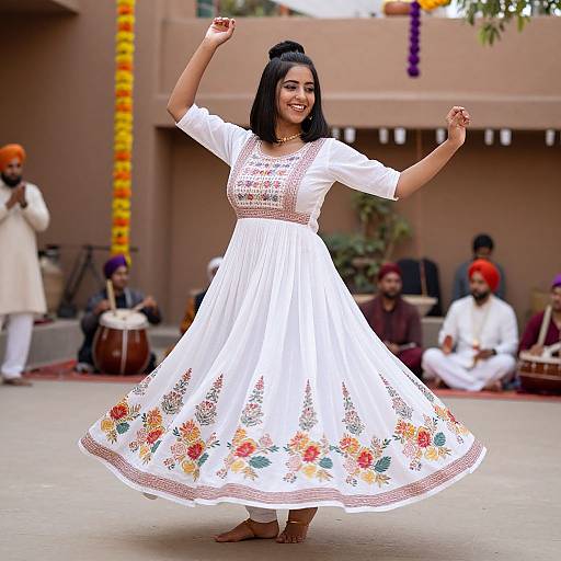 Photograph of a smiling South Asian woman dancing in a white traditional Punjabi dress with floral embroidery, arms raised, in a courtyard with musicians and colorful