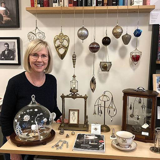 Photograph of smiling blonde woman with glasses, black sweater, in jewelry shop, displaying intricate hanging ornaments, teapot, glass dome, on wooden table