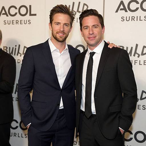 Photograph of two men in black suits and white shirts, standing together with one arm around the other, in front of a 