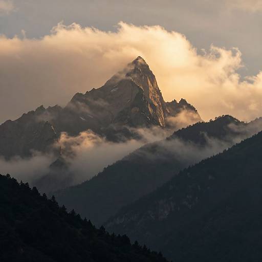 Photograph of a rugged mountain peak bathed in golden sunlight, partially obscured by clouds and dark forested hills in the foreground.