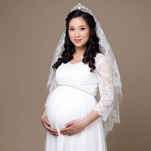 Photograph of a pregnant Asian bride in a white lace wedding dress and veil, gently cradling her belly, smiling, against a beige background.