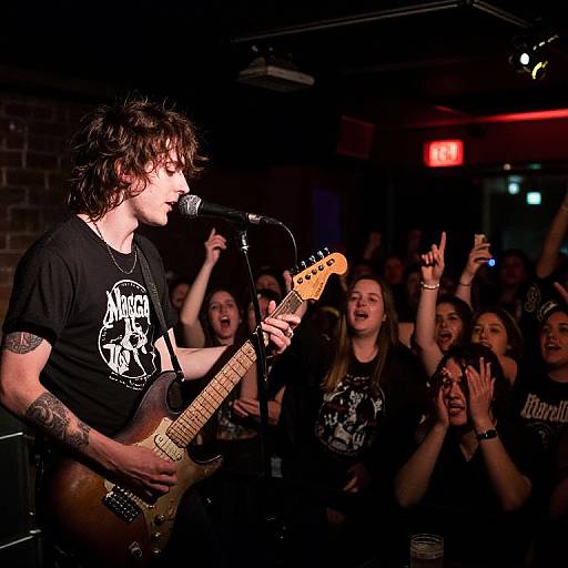 Photograph of a male bassist with curly hair, black t-shirt, and tattoos, performing on stage to an enthusiastic, cheering crowd.