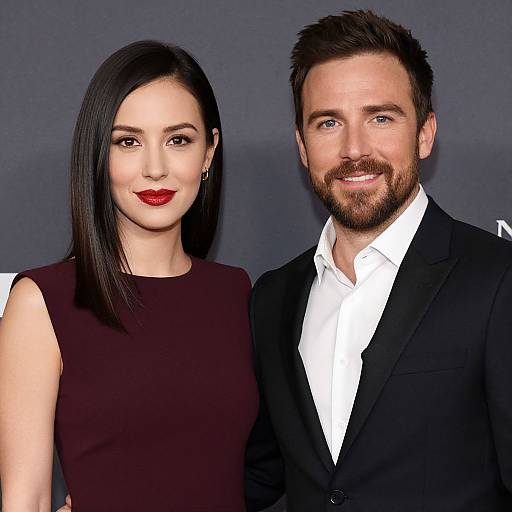 Photograph of a smiling couple; woman with straight black hair, red lipstick, sleeveless dark dress, man with brown beard, black suit, white