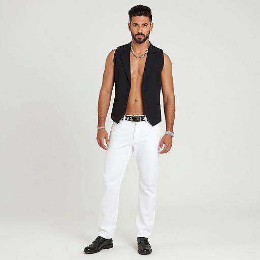 Photograph of a bearded man with short dark hair, wearing a black sleeveless vest, white pants, black shoes, and jewelry, standing against