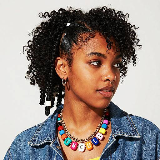 Photograph of a young Black woman with curly hair, wearing a denim shirt and colorful bead necklace, looking to the side. White background.