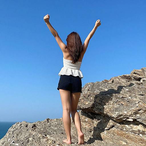 Photograph of a woman with long brown hair, white strap top, black shorts, and barefoot, standing on rocky cliff with arms raised, against