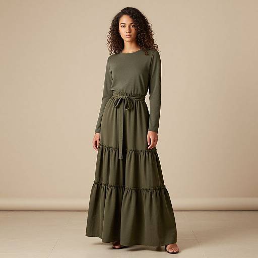 Photograph of a curly-haired woman in a long, olive-green, three-tiered dress with long sleeves, standing against a plain beige background.