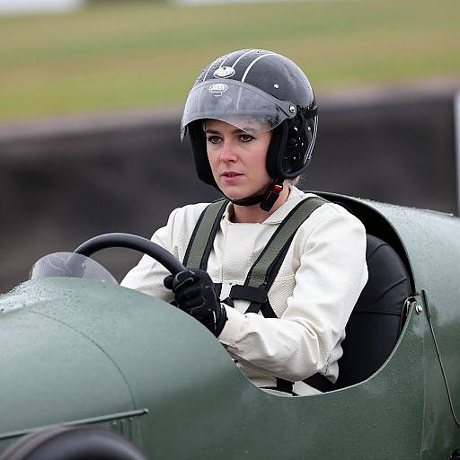 Photograph of a serious young woman with light skin and black helmet, wearing white shirt and black gloves, driving a green vintage aircraft.
