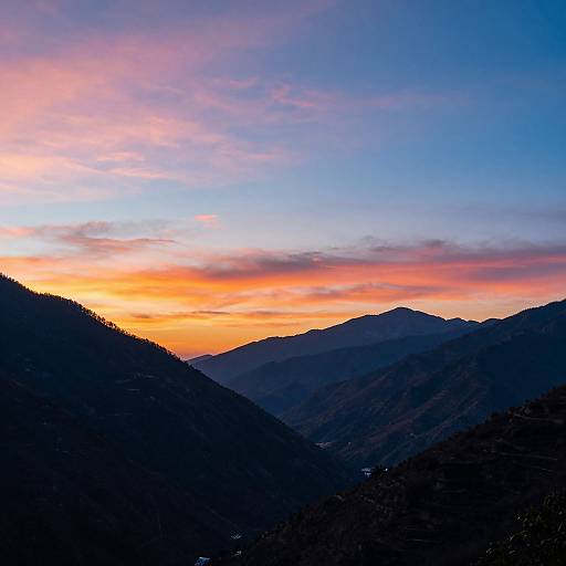 Photograph of a sunset over mountainous landscape, with vibrant orange and pink sky, silhouetted dark mountains, and layered shadows.