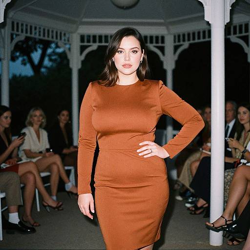 Photograph of a confident woman with dark hair in a long-sleeve, form-fitting, rust-colored dress, standing in front of an outdoor