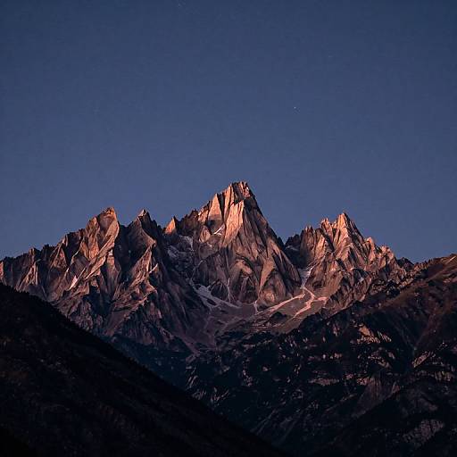 Photograph of a rugged, sunlit mountain peak bathed in pink and purple hues, with dark shadows below and a clear, deep blue night sky