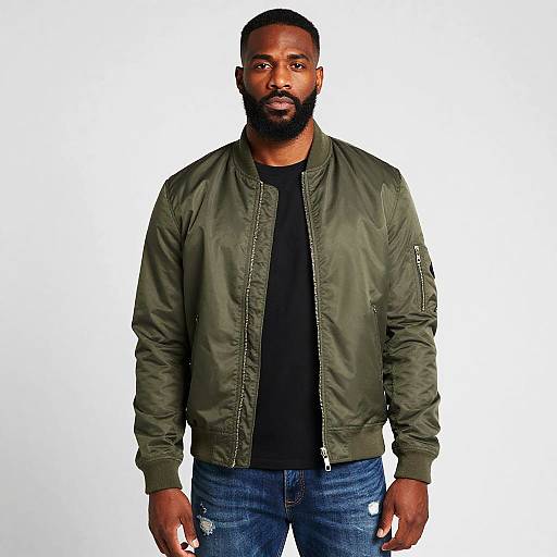Photograph of a muscular, bearded Black man with short hair, wearing a green bomber jacket, black shirt, and blue jeans, standing against a