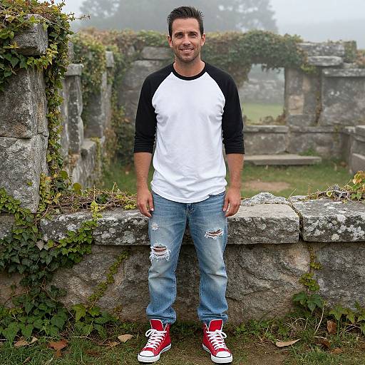 Photograph of a smiling man with short dark hair, wearing a white and black raglan shirt, ripped jeans, and red sneakers, standing against a