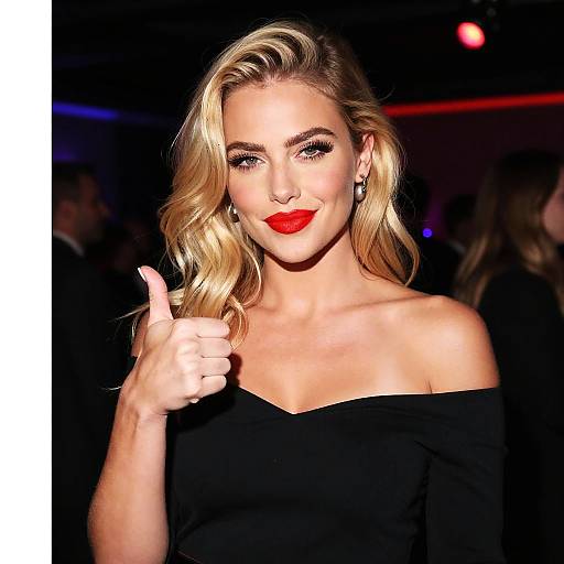 Photograph of a blonde woman with wavy hair, red lipstick, and black off-shoulder top, giving a thumbs-up in a dimly