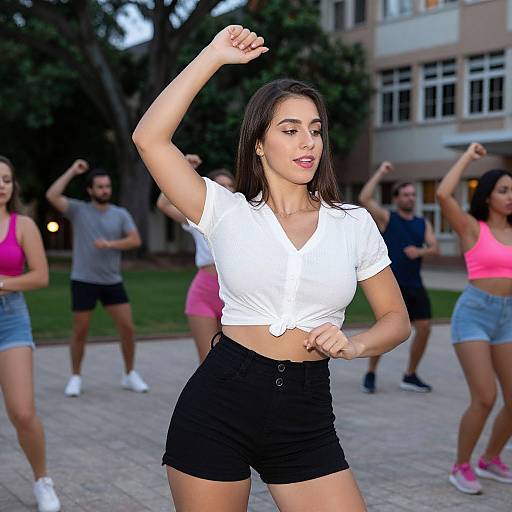 Photograph of a young woman with long brown hair, wearing a white tied-up crop top and black shorts, dancing outdoors with friends in a park.