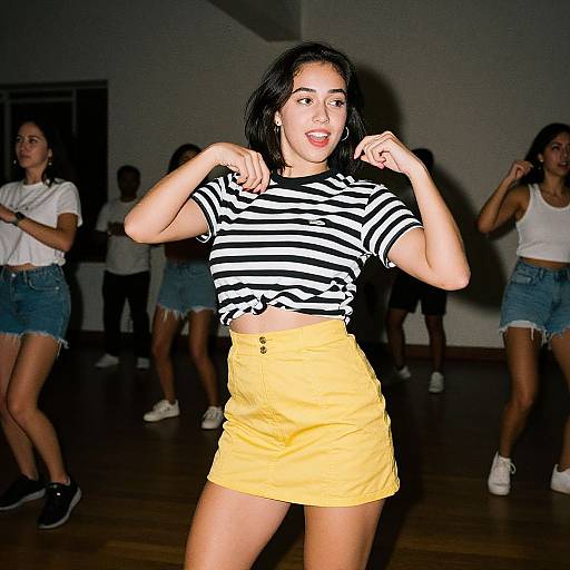 Photograph of a young woman with black hair, wearing a black-and-white striped shirt and yellow shorts, dancing in a dimly lit room with other