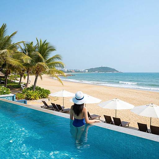 Photograph of a woman in a blue swimsuit and white sunhat, standing in a blue infinity pool, facing a sunny, palm-lined beach with
