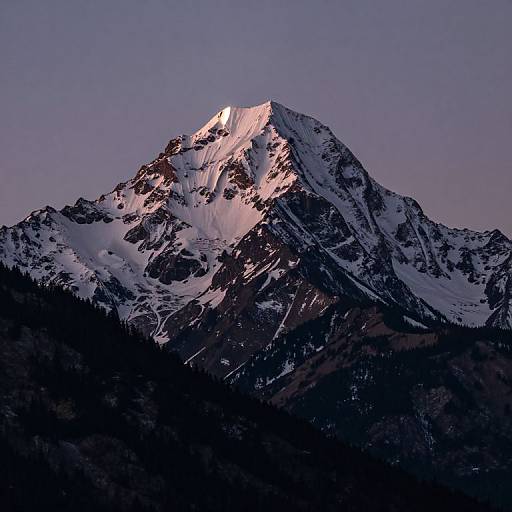 Photograph of a snow-capped mountain peak illuminated by sunset light, with dark rocky slopes and a clear purple-blue sky.