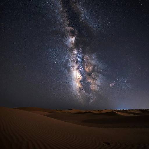 Photograph of a starry night sky over a desert, with the Milky Way galaxy prominently visible, illuminating dark, rippled sand dunes below