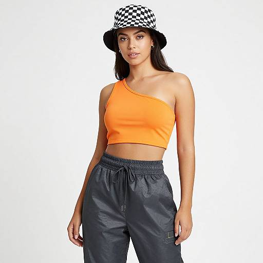 Photograph of a young woman with light brown skin, wearing an orange one-shoulder crop top, black checkered bucket hat, and dark blue
