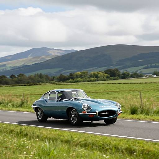 Photograph of a blue vintage sports car driving on a narrow road through a lush green countryside with rolling hills and mountains in the background.