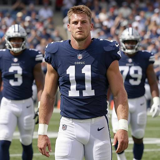 Photograph of muscular, blonde male football player in navy jersey number 11, white pants, standing on field with blurred teammates.