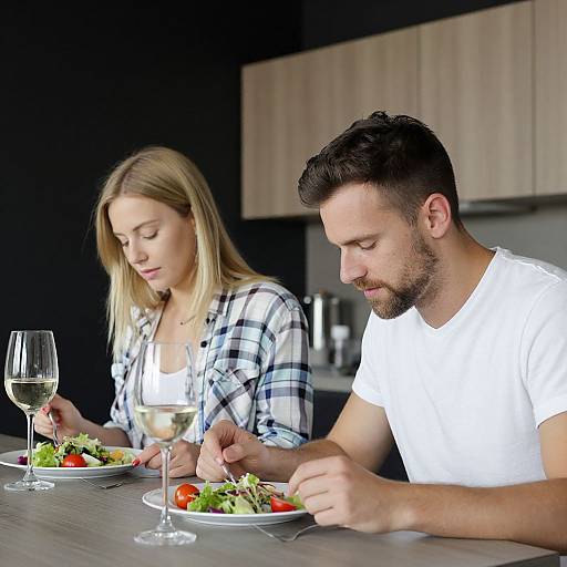 Photograph of a blonde woman and a bearded man in a modern kitchen, eating salads with wine glasses, focused on their plates.