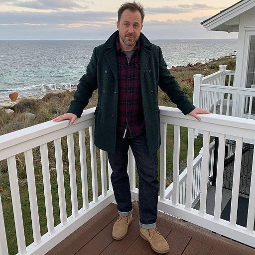 Photograph of a bearded man with short dark hair, wearing a black coat, red plaid shirt, black pants, and tan boots, standing