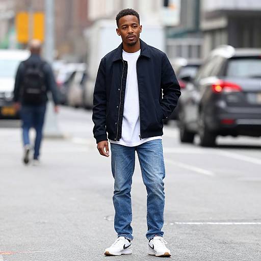Photograph of a Black man with short hair, black jacket, white shirt, blue jeans, and white sneakers, walking on a busy urban street.