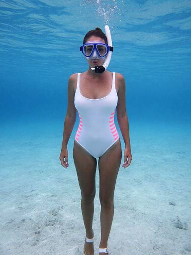 Photograph of a woman underwater, wearing a white one-piece swimsuit with pink stripes, blue snorkel mask, and white fins, standing on a