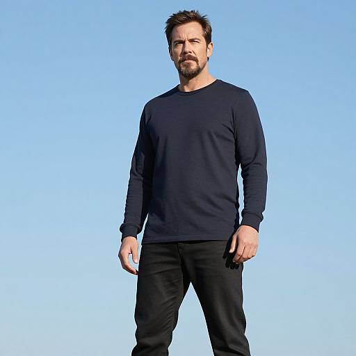 Photograph of a bearded man with short brown hair, wearing a black long-sleeve shirt and black pants, standing against a bright blue background