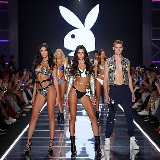 Photograph of a fashion runway show featuring three models: two women in sparkly bikinis and one man in an open, patterned vest and pants
