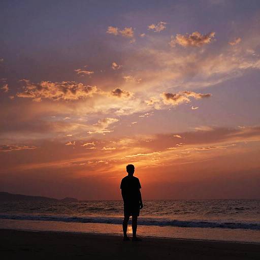 Silhouetted man standing on beach at sunset, colorful sky with orange, pink, and purple clouds, waves gently touching shore. Photograph.