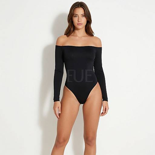 Photograph of a young woman with long brown hair, wearing an off-shoulder black long-sleeve leotard, standing against a plain