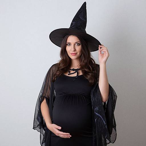 Photograph of a pregnant woman with long brown hair, wearing a black witch hat, black dress, and sheer black shawl, touching her hat,
