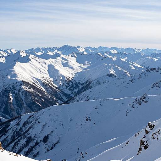 Photograph of a vast, snow-covered mountain range under a clear blue sky, with sunlight highlighting jagged peaks and dark rocky outcrops.