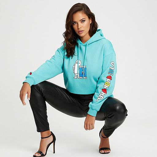 Photograph of a confident woman with wavy brown hair, squatting, wearing a light blue hoodie with colorful graphics, black pants, and black high