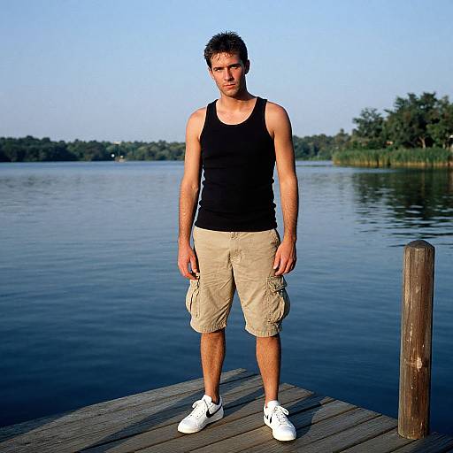 Photograph of a muscular, short-haired man in a black tank top and beige cargo shorts, standing on a wooden dock by a calm lake under a
